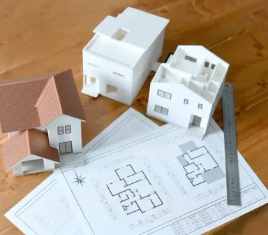 Weiße Architekturmodelle von Einfamilienhäusern neben einem Grundriss-Bauplan und einem Lineal auf einem Holztisch